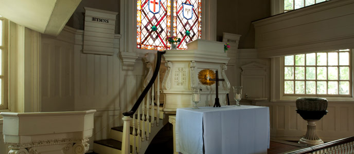 Color photo of the interior at Gloria Dei Church, showing a cross standing on a white cloth covered table, with a large stained glass window behind. Color photo of the interior at Gloria Dei Church, showing a cross standing on a white cloth covered table, with a large stained glass window behind.