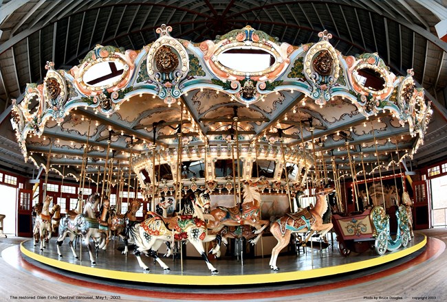Photograph of a historic carousel with brightly painted carousel horses, one deer and the back of a carousel chariot in front.