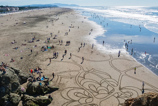 View of ocean beach crowds on a beautiful day, including someone creating sand art with a rake, from an overlook at the beach's north end