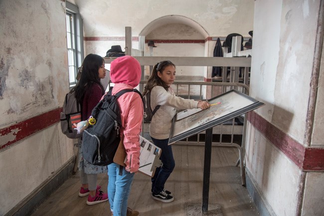 Three students with worksheets read an exhibit sign inside Fort Point.