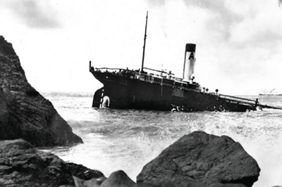 Large ship half submerged in water with surf and rocks nearby.
