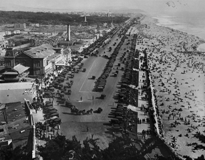 Aerial of crowded beach and adjacent highway filled with old-fashioned cars.