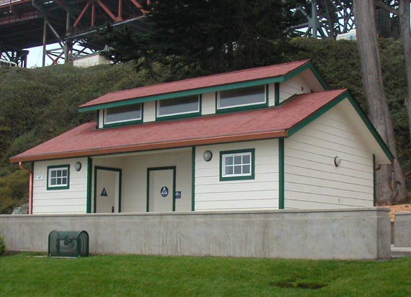 White restroom building with red roof surrounded by trees under a bridge.