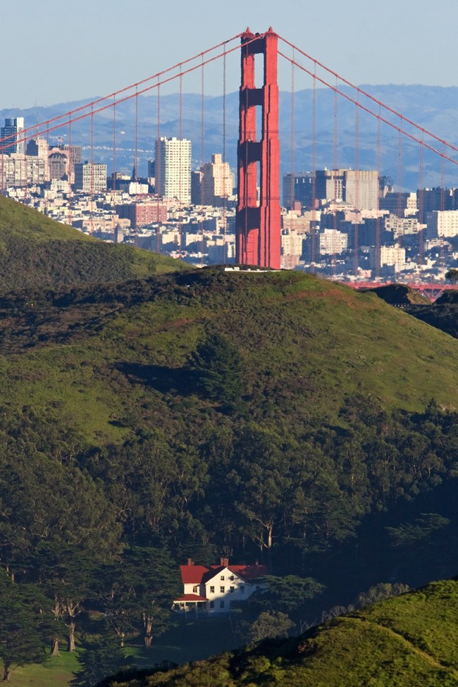 A white building with a red roof is nestled within the rolling green Marin Headlands. The Golden Gate Bridge is in the background.