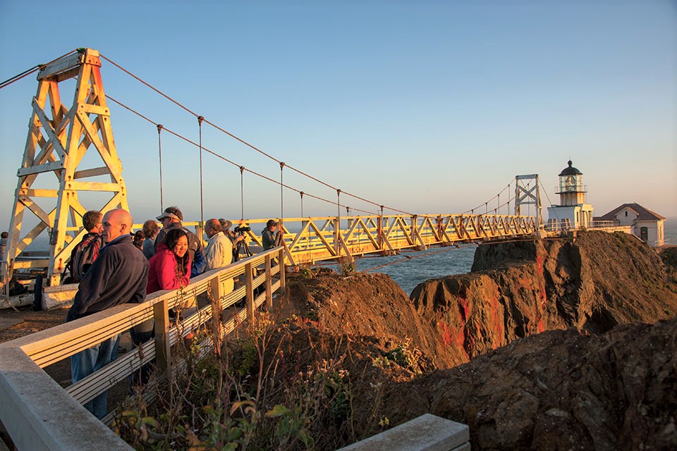 People gather at the bridge to Point Bonita Lighthouse as the sun sets with the ocean in the background