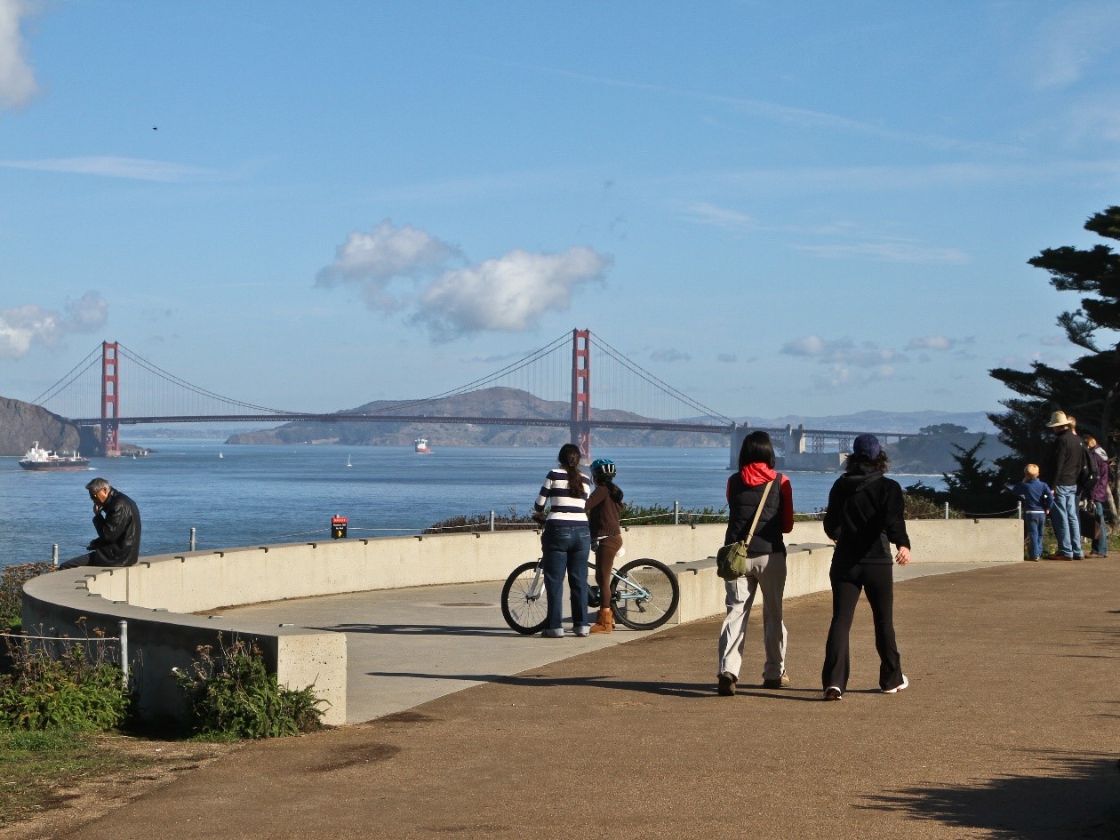 Visitors stand at a lookout at Lands End facing the Golden Gate Bridge which sits below a blue sky with clouds.
