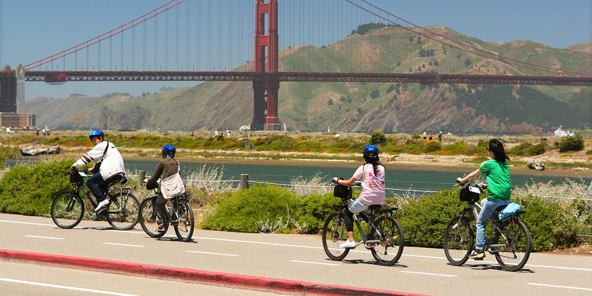 Four people pedal on bicycles along a path in front of the lagoon at Crissy Field as the Golden Gate Bridge stands in the background.
