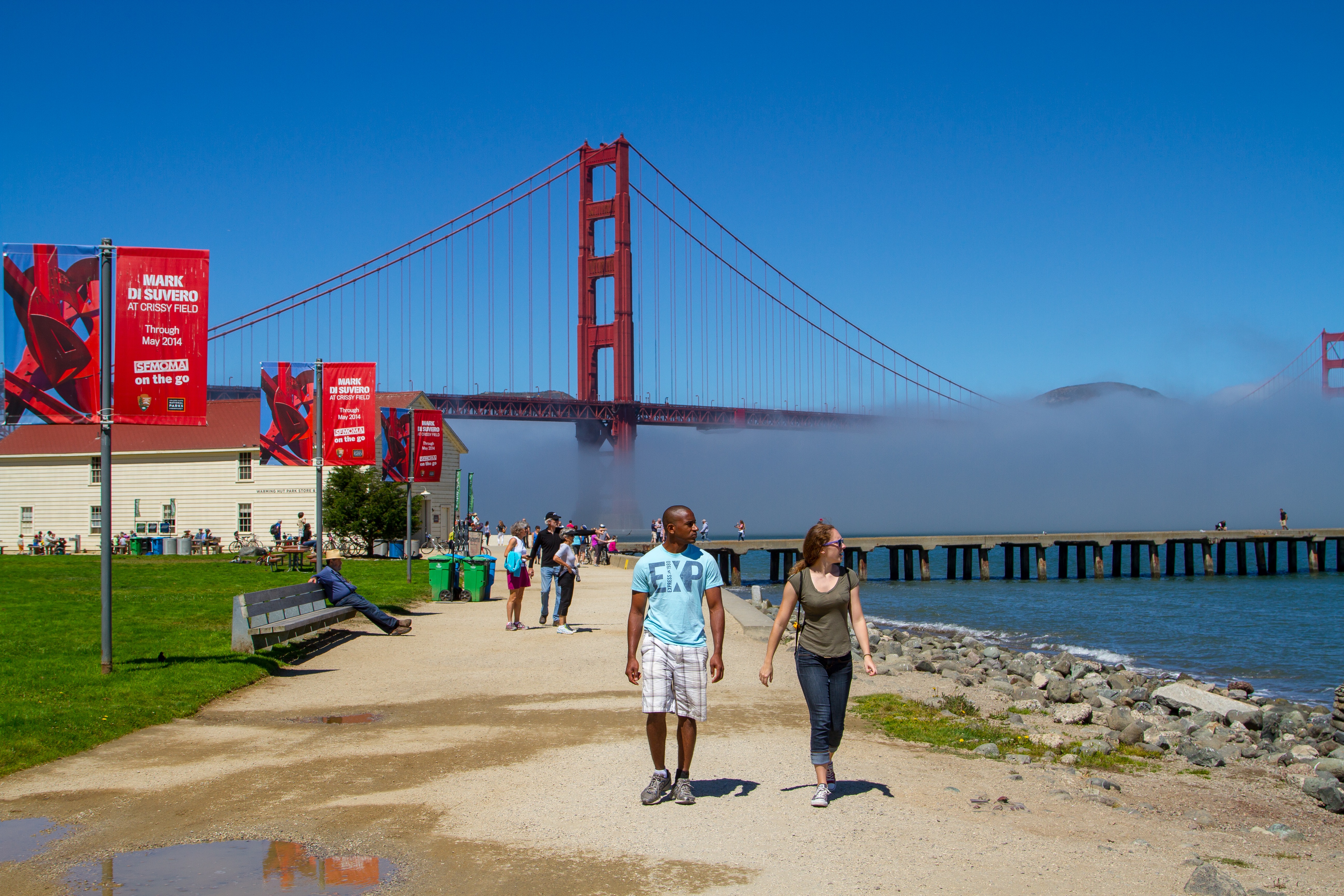 People walk along San Francisco Bay on a bright blue day with the Golden Gate Bridge and fog in the background. The white Warming Hut and red banners are left of the trail.