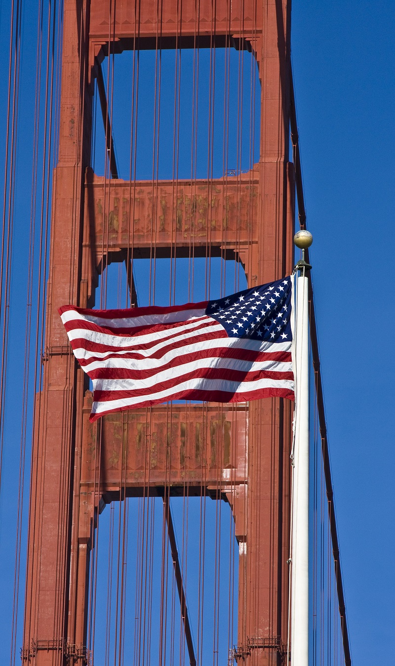 American flag at full staff with a tower and cables of the Golden Gate Bridge in the background.