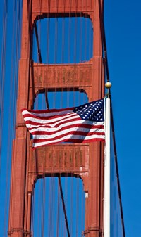 American flag at full staff with a tower and cables of the Golden Gate Bridge in the background.
