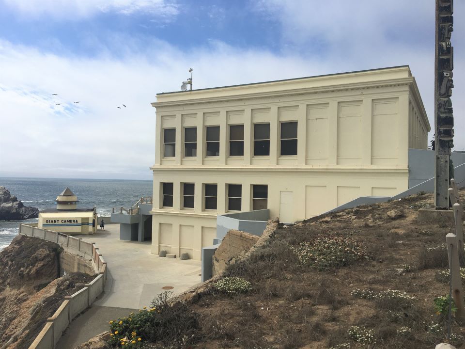 A side view of a beige building that sits on a cliffside with the ocean and rocks in the background.