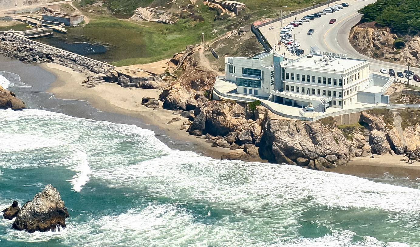 Curved road leading up to a white three floor building on the edge of a cliff. Cars parked diagonal along the left and right side of the building. Below the rocky cliff are crashing waves.