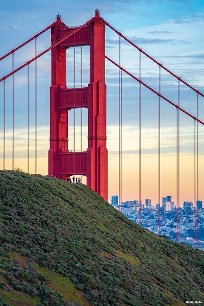 People standing on a lush green hill with one of the Golden Gate Bridge towers and its support cables beside the hill. The city of San Francisco in the background.