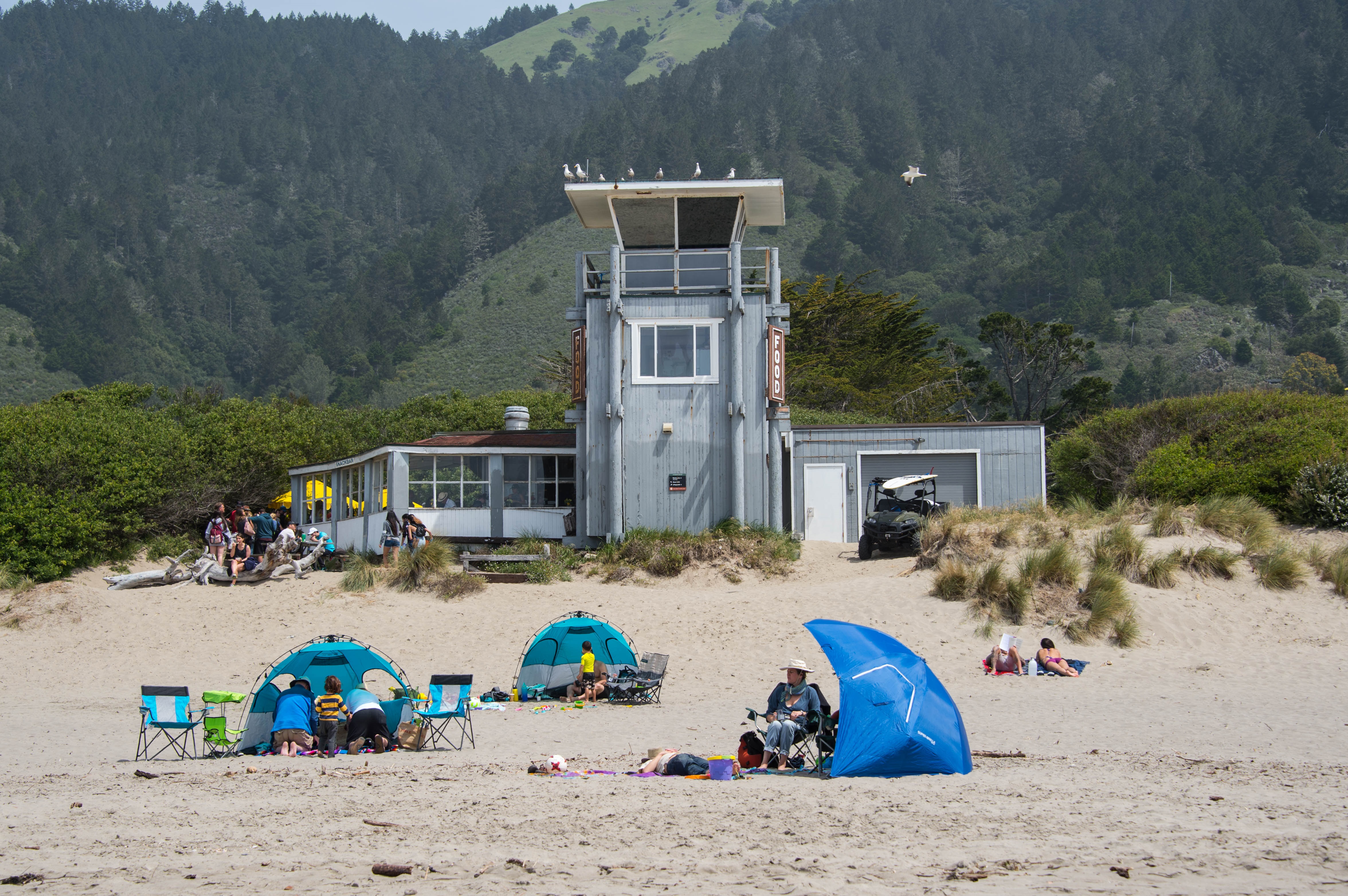 Two-level tower with people and tents in the front sitting on sand and green hillside in the background. Two-level tower with people and tents in the front sitting on sand and green hillside in the background.