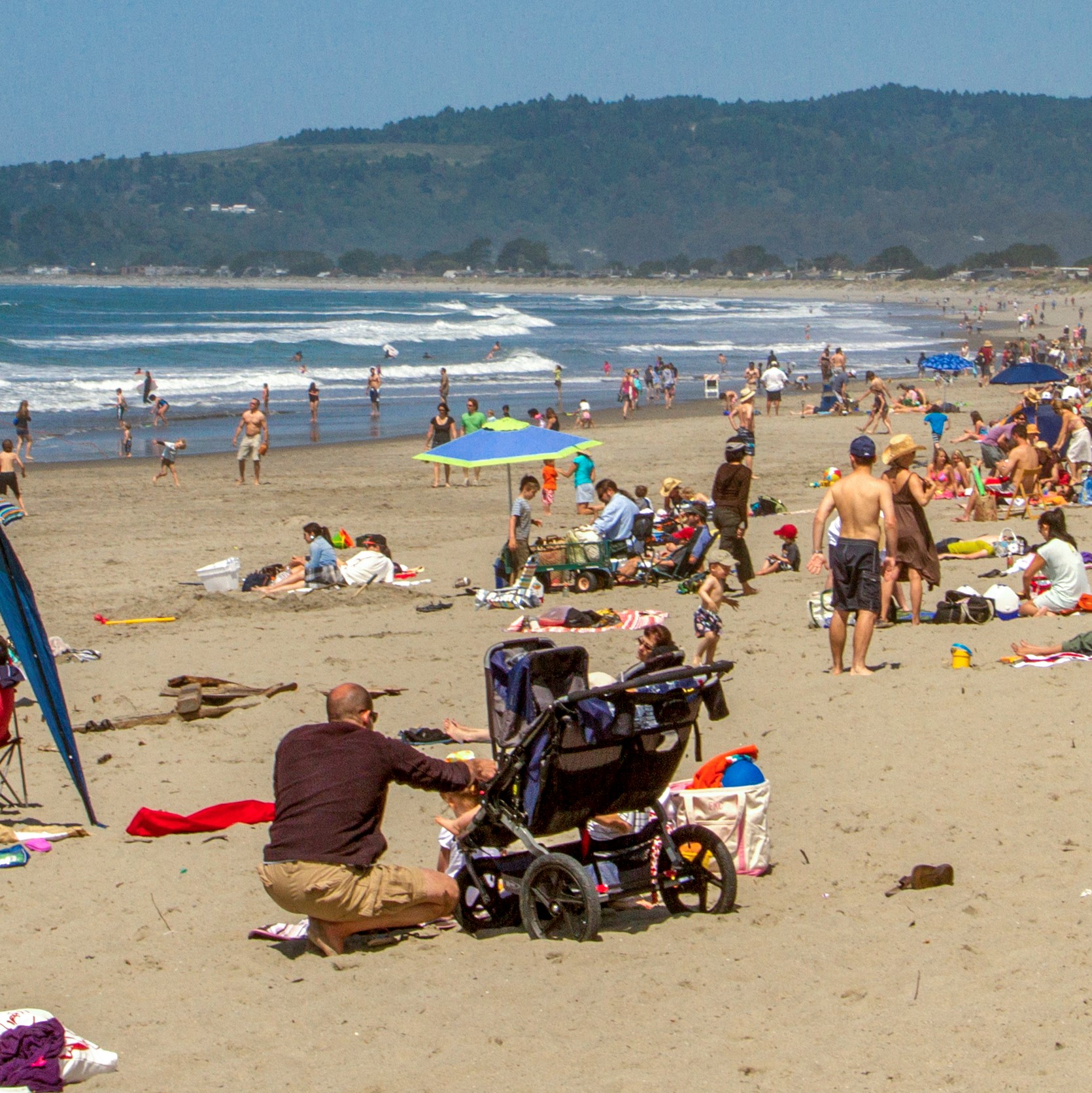 Under a blue sky visitors line a sandy beach as waves come toward the shore.