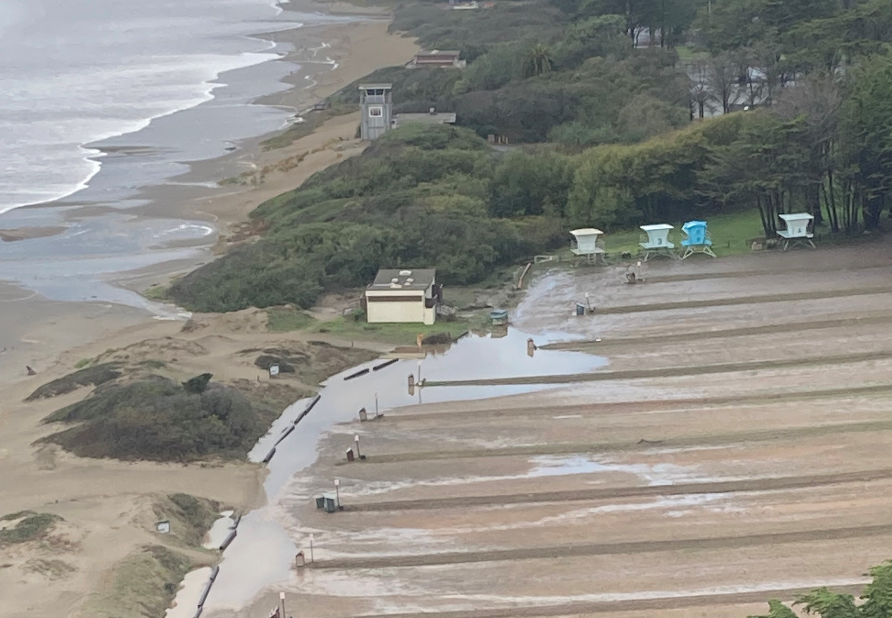Color photo of Stinson Beach south parking lot with standing water on left side of picture. Pacific Ocean visible at top left and restrooms and lifeguard towers visible along left and top edges of parking lot. Photo taken after October 2021 rainstorm.
