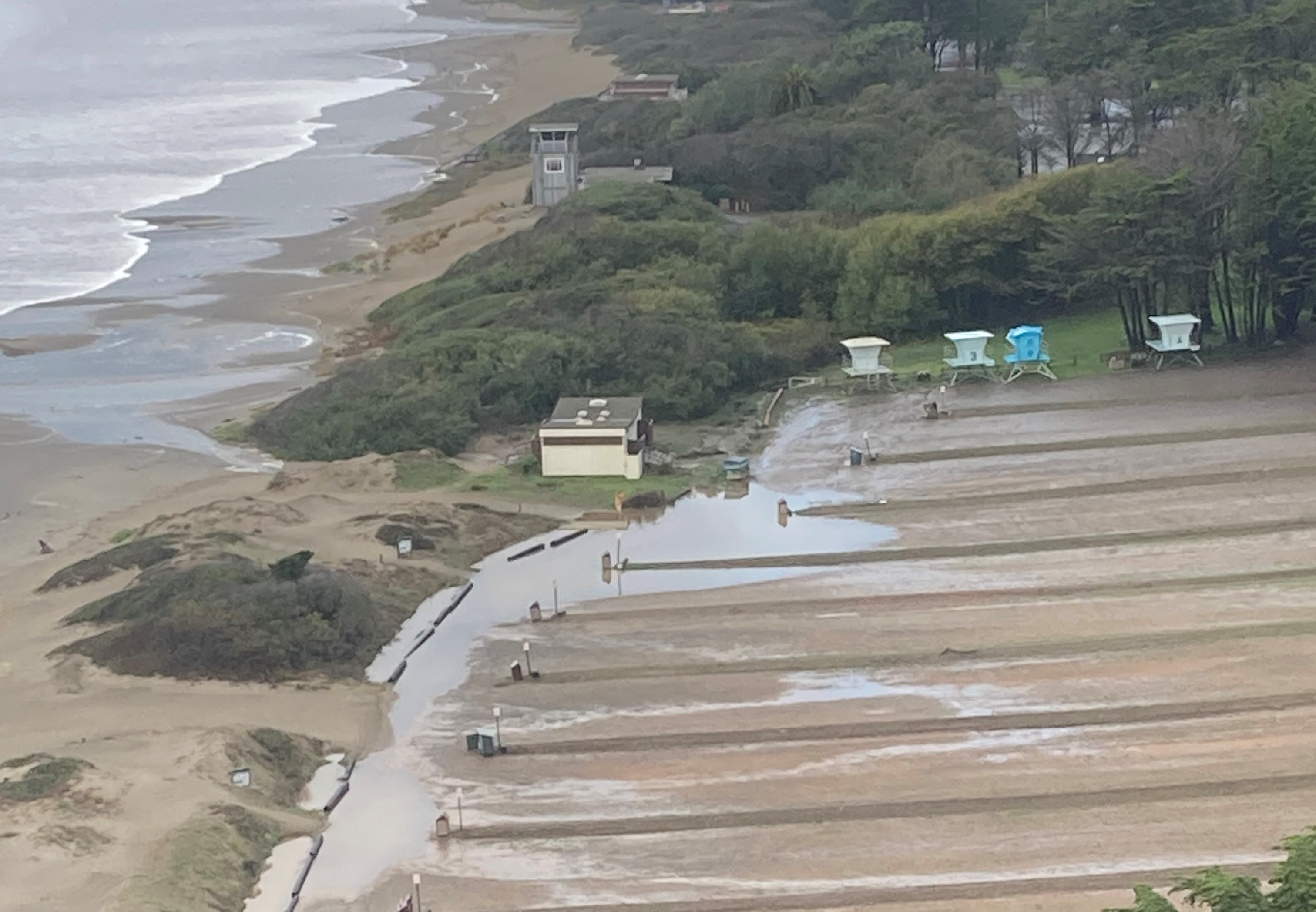 Stinson South Lot NPS Xave Agnew Oct 2021 Color photo of Stinson Beach south parking lot with standing water on left side of picture. Pacific Ocean visible at top left and restrooms and lifeguard towers visible along left and top edges of parking lot.