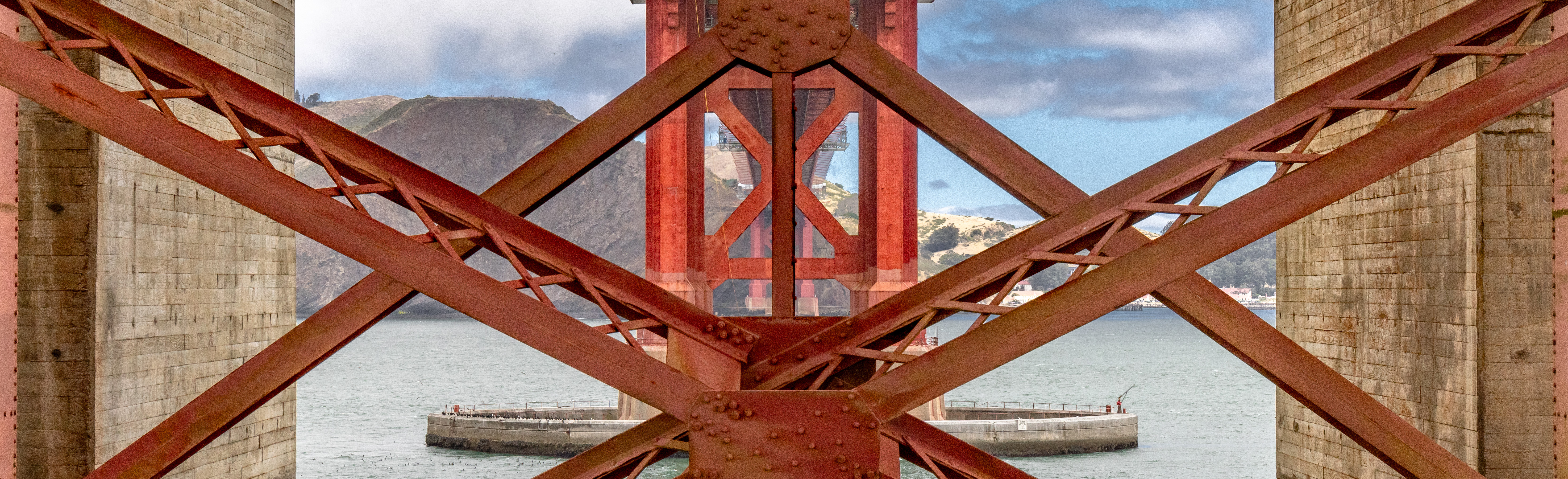 Color photo of international orange Golden Gate Bridge trusses with green coastline in background.