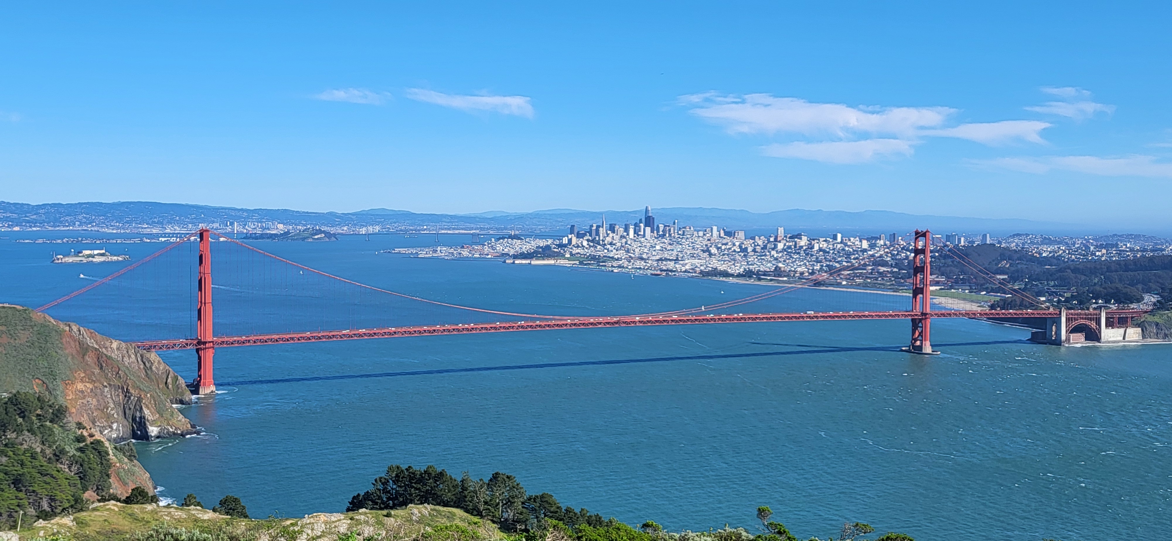 Golden Gate Bridge spanning San Francisco Bay with green hillsides of Marin Headlands in foreground and City of San Francisco in background.