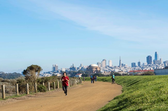 A man jogs along a wide dirt path. A city skyline is visible in the distance