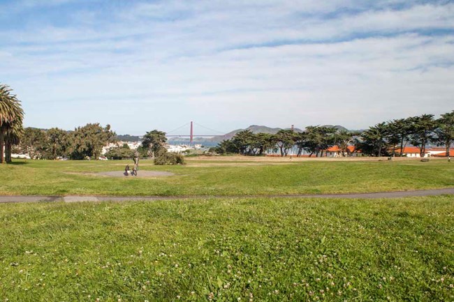 A paved path runs across a grass lawn. A large orange suspension bridge can be seen in the distance.