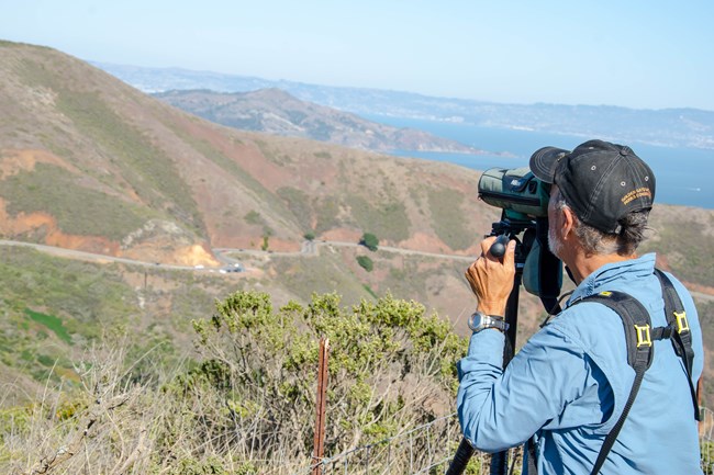 A person with binoculars looks out into a valley surrounded by mountains