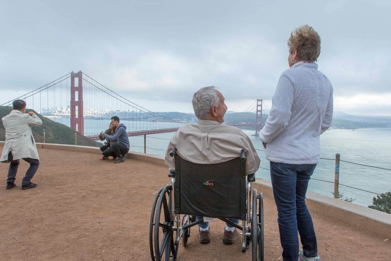 Man in a wheelchair besides a standing women overlooking an orange bridge