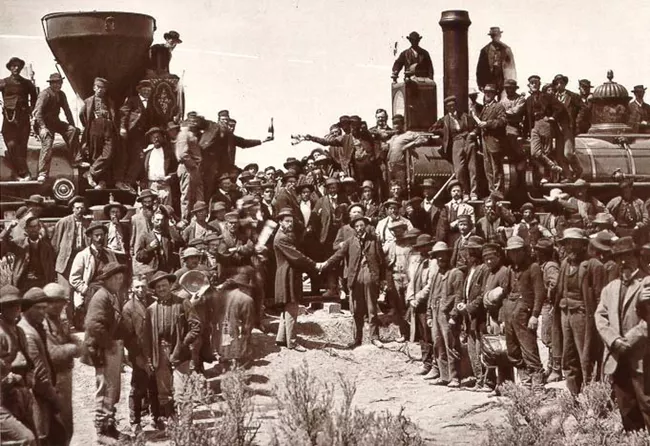 Groups of White workers gathered between and on top of two large locomotives at the Golden Spike ceremony. Two men one the front shake hands.