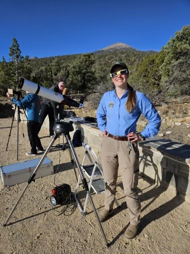 A color photo of a smiling woman standing next to a telescope during the day. She wears a bright blue shirt with an emblem near the shoulder, along with a hat and eclipse glasses with very dark lenses. The telescope is a solar scope pointed to the left.