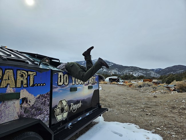 A color photo of a person with their torso inside of a large dumpster. Their legs flail in the air comedicaly with a snowy mountain in the distance