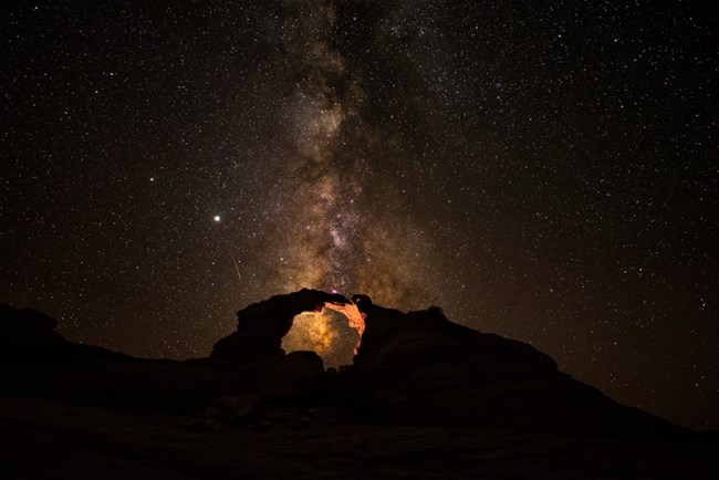 A color photo of the milky way stretching above a rock arch lit from within