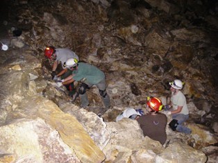 Four individuals with brightly colored helmets sit on a field of boulders. Two are sitting while two others inspect the rock at their feet, reaching out with their hands to examine it.