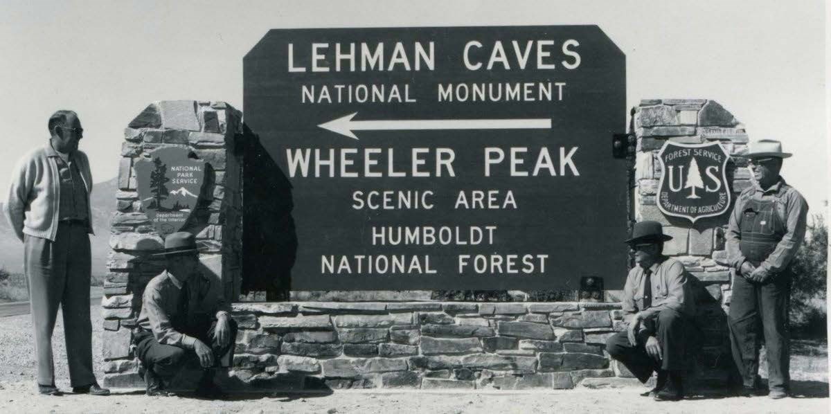 Black and White photo of a total of 4 park staff members standing on both sides of the Park Entrance Sign. The sign reads "Lehman Caves National Monument, Wheeler Peak Scenic Area, Humboldt National Forest".