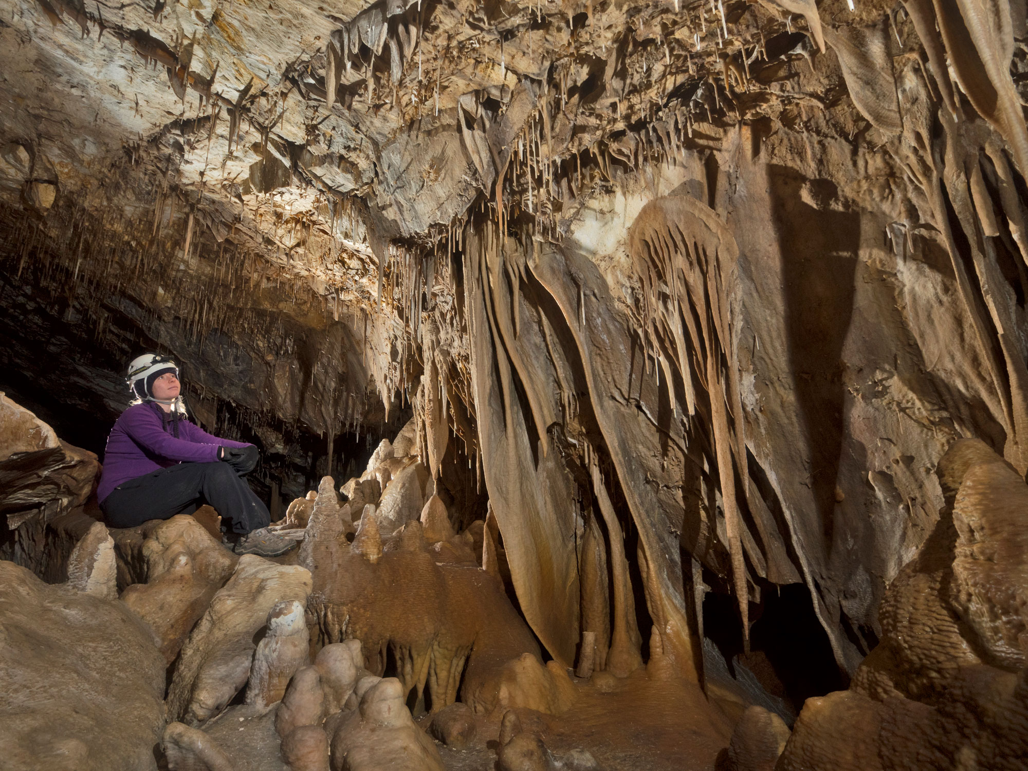 PR---Additional-Lehman-Caves-Tours---Wild-Cave-Tour---Talus-Room-Web--Copyright-David-Bunnell A caver sits on a rock shining her headlamp onto drapery formations cascading down a rock wall.