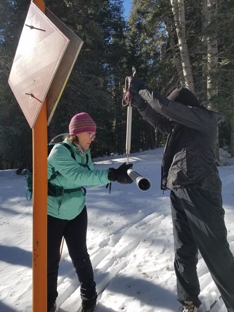 one person holding a scale, the other balancing a tube in a wintery scene with an orange snow survey sign in the foreground