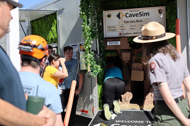 A group of high school students, some wearing safety helmets, watches a friend begin crawling through the Cave Simulator trailer.
