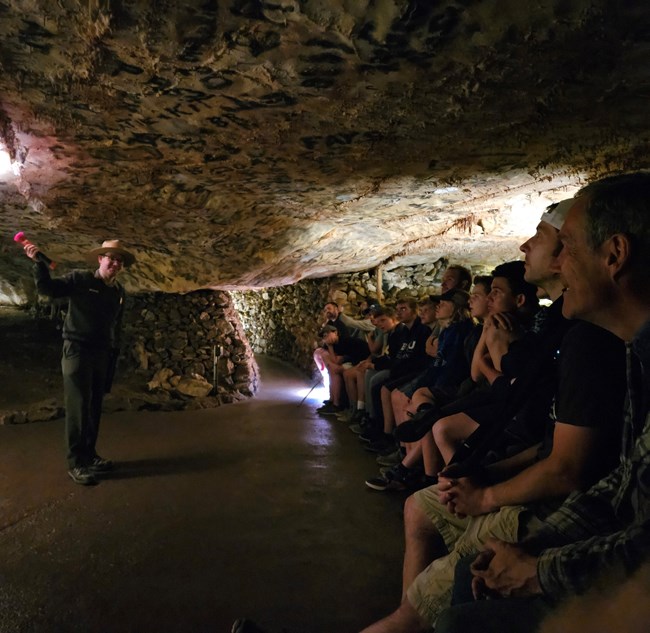 A park ranger stands to one side of a cave room. On the other side is a row of about twenty seated people watching intently as the ranger speaks.