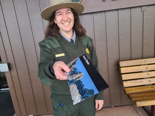 A female park ranger in uniform holds a brochure titled "Great Basin" toward the camera, with "Lehman Caves Visitor Center" sign behind her.
