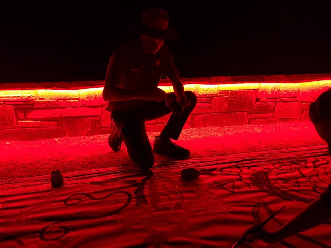 A park ranger kneels over a canvas splayed out on the ground. A small rock wall with an underglow of red light illuminates the canvas and ranger.
