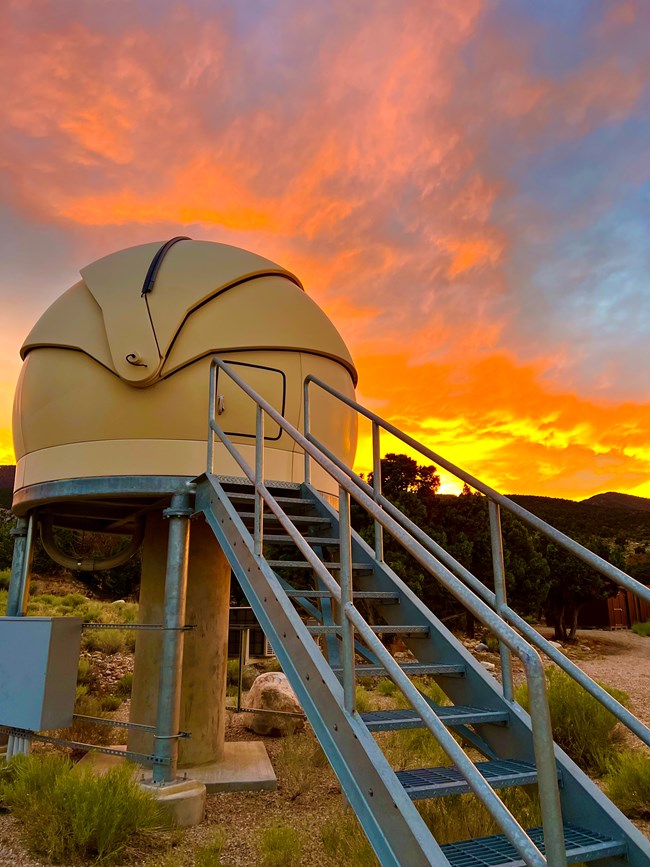 Silver stairs lead several feet into the air to a white dome suspended on a metal and concrete pillar. A vivid orange sunset lights the distant mountains.