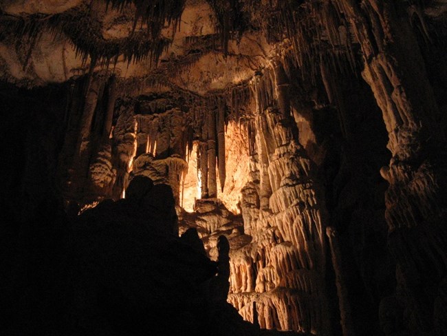 A dimly lit, rocky room filled with various cave formations. The gold light illuminates tan and white stalactites, stalagmites, and other smaller formations.
