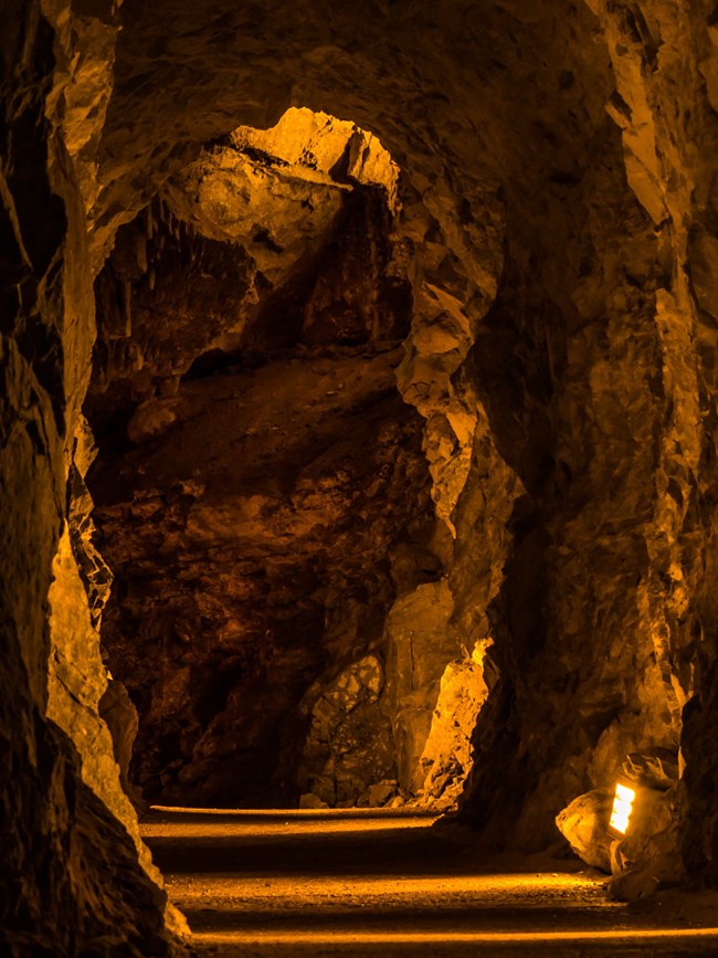 An amber lit rocky passageway leads deeper into a cave. A flat floor with amber lights only inches up the walls beckon visitors deeper.
