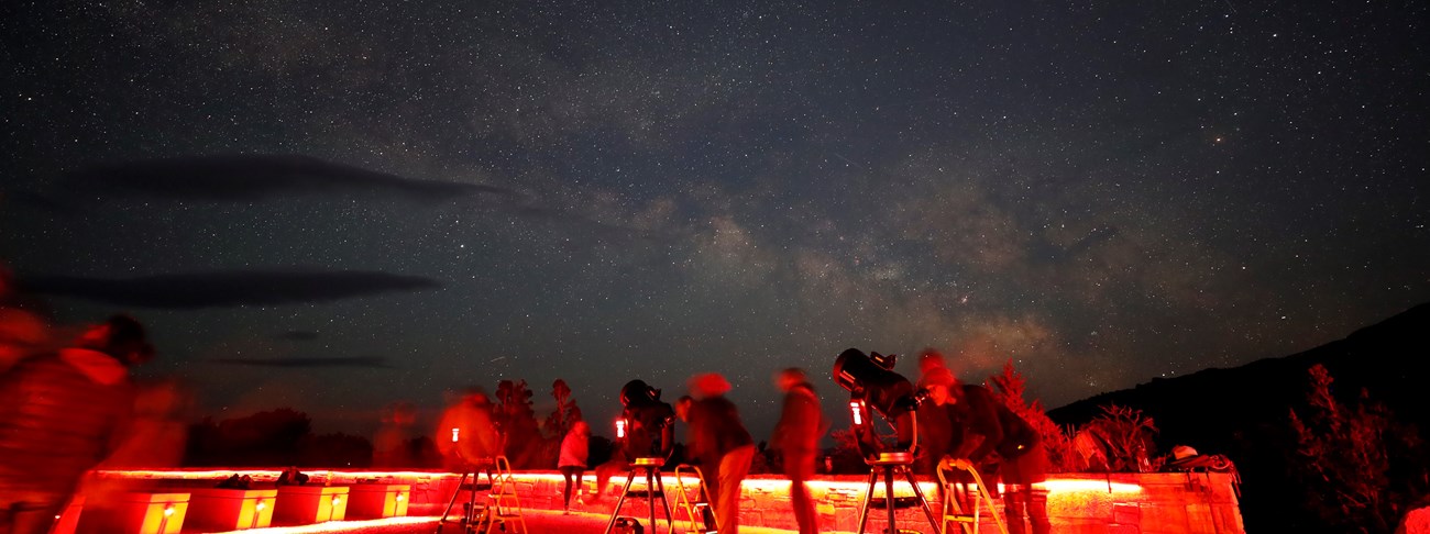 Three telescopes with shadowy figures due to long exposure stand in front of a colorful milky way arcing across the star filled night sky