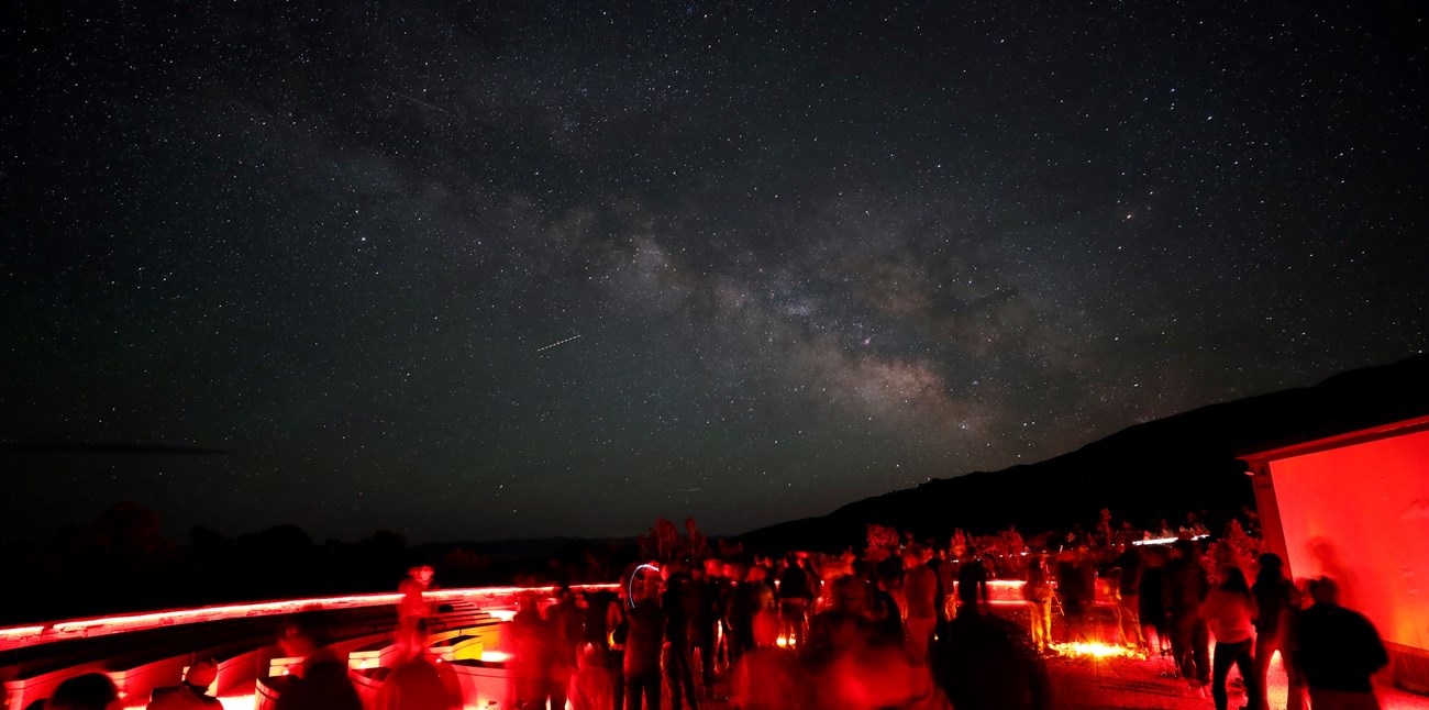 Red lights fill a dark amphitheater with blurry figures as the colorful milky way arcs across the night sky