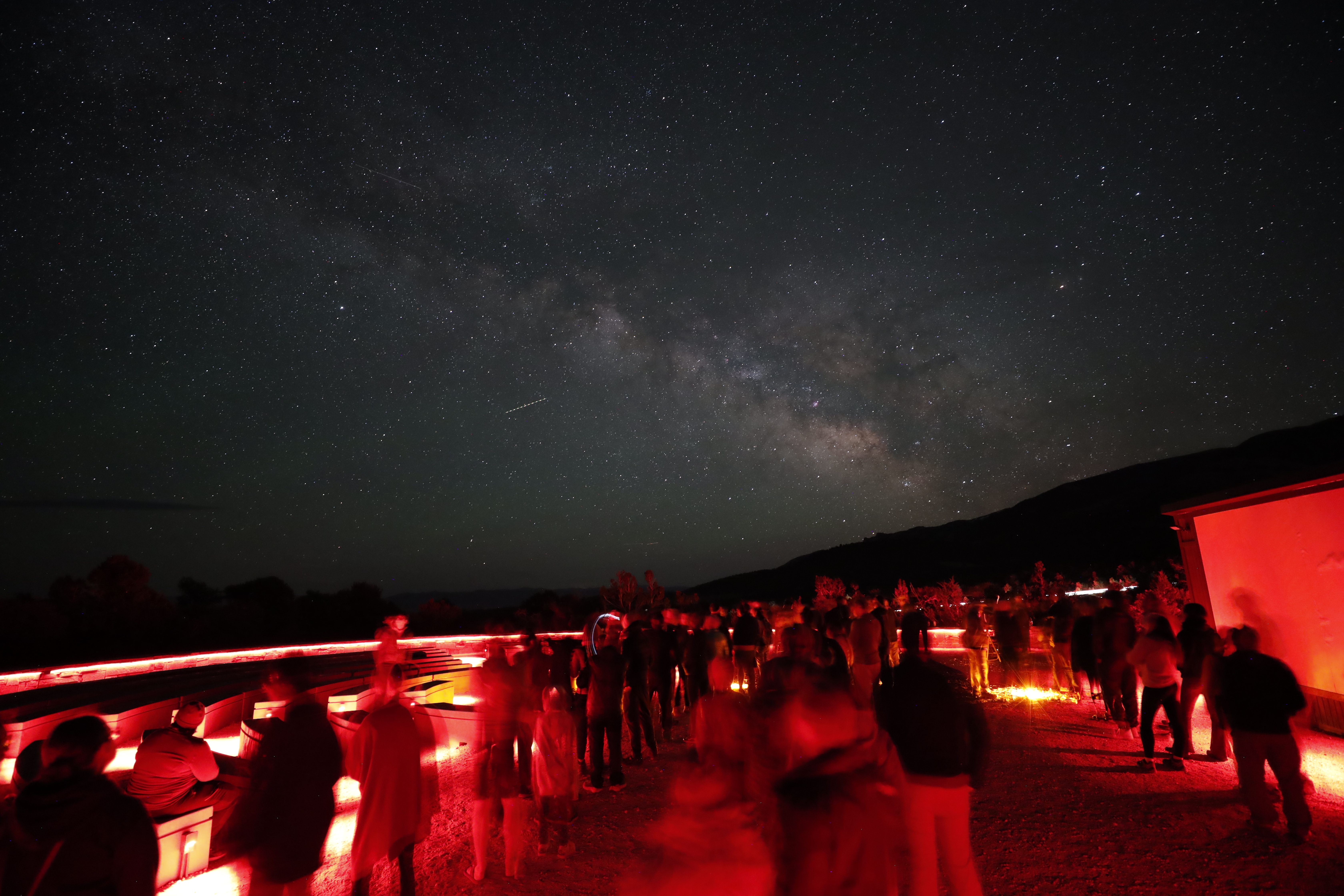 Red lights fill a dark amphitheater with blurry figures as the colorful milky way arcs across the night sky