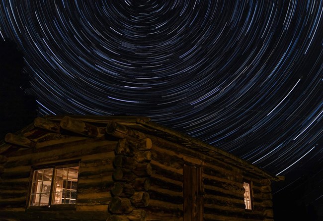 A wooden cabin with light eminating from two windows lies beneath a swirling image of the night sky. Long exposure photography creates trails of the stars as the earth rotates. The swirls center around the unmoving north star Polaris.