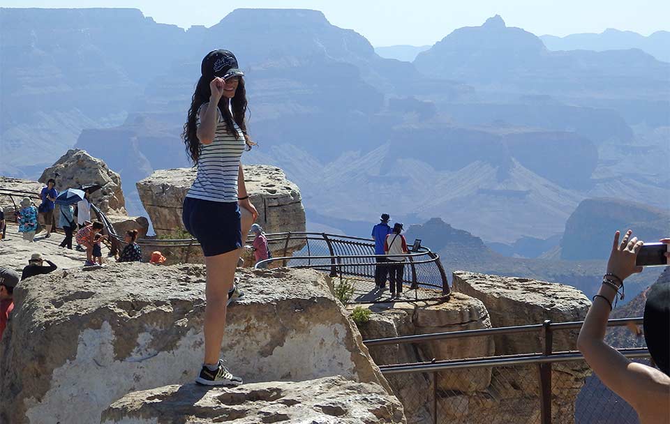 Two women at scenic Grand Canyon overlook with railings. One woman is standing on a rock and striking a pose. The second woman is on the far right and is taking a photo with her phone of the first woman.