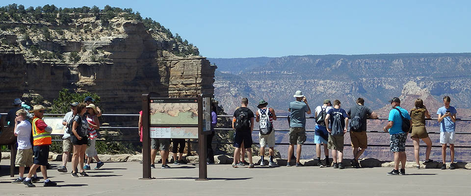 A number of park visitors dressed in summer attire are standing at a metal guardrail and looking out at Grand Canyon.