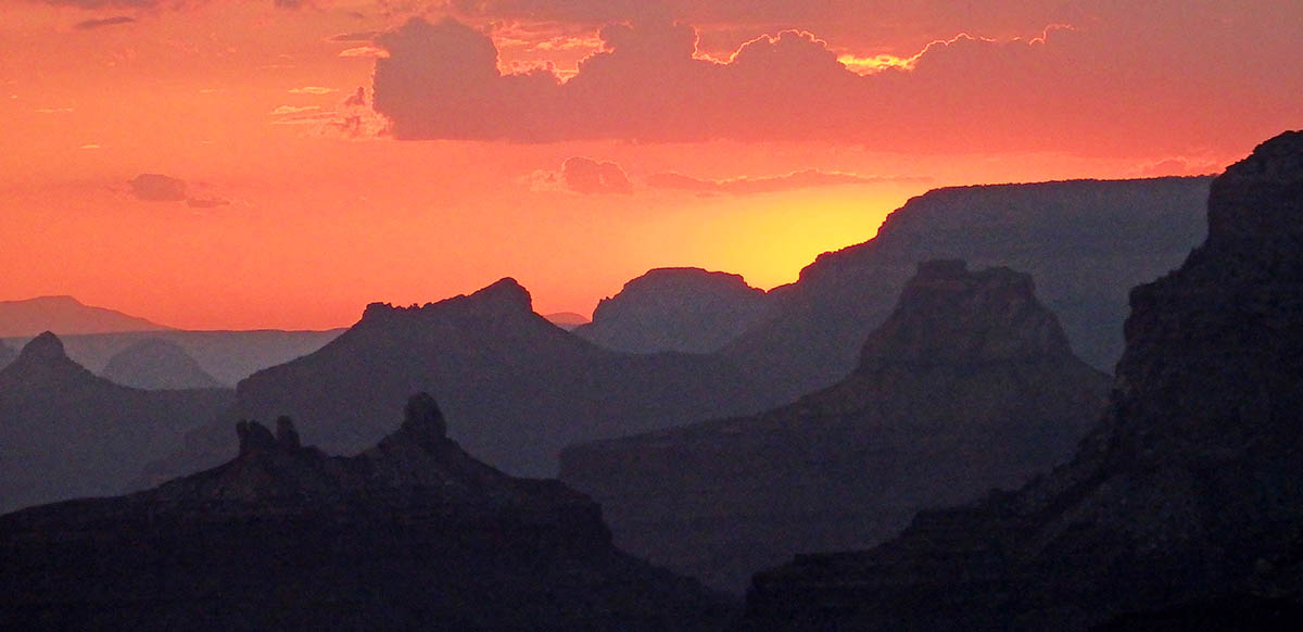 Looking west at sunset through a number of silhouettes of buttes and ridge lines within Grand Canyon. The sky, clouds, and everything in view is bathed in reddish-orange light. Looking west at sunset through a number of silhouettes of buttes and ridge lines within Grand Canyon. The sky, clouds, and everything in view is bathed in reddish-orange light.
