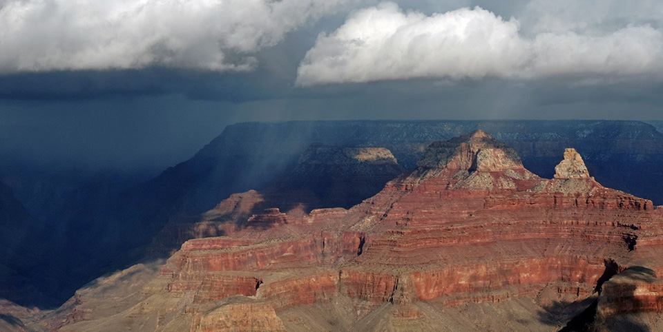 White summer thunderstorm clouds overhead are shadowing out the North Rim in the background. Bright sunlight is falling onto white capped red stone temples in the foreground. 